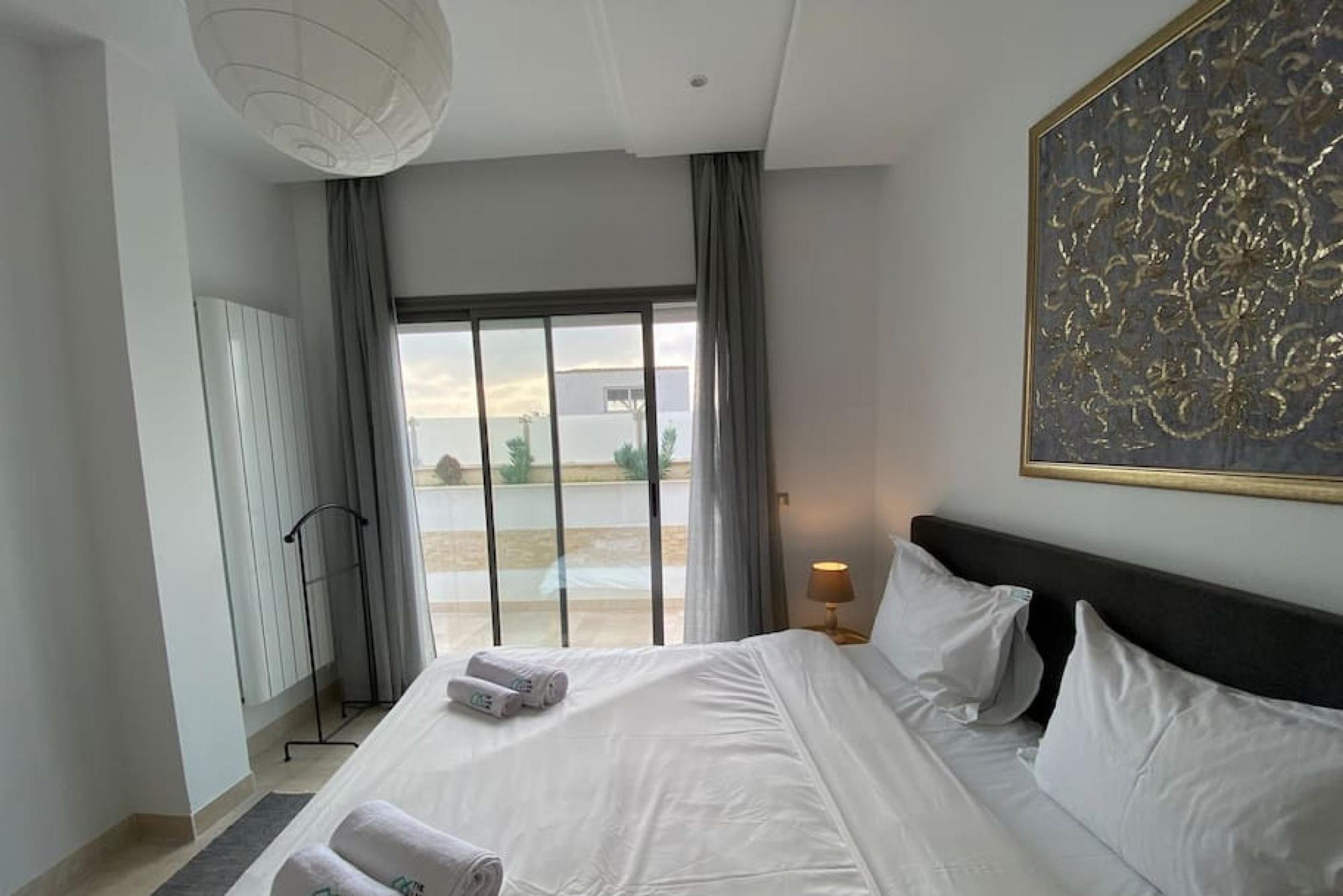 A modern hotel room with a large bed, a view through the window, and a painting on the wall.