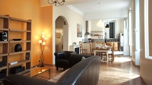 Un salon avec un canapé, une table basse et une bibliothèque. On y trouve également un coin repas avec une table et des chaises. La cuisine est visible au fond.