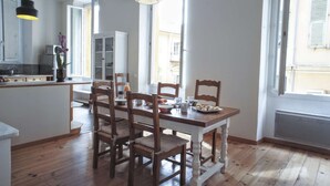 Un coin repas avec une table et des chaises en bois, une cuisine à l’arrière-plan et de grandes fenêtres laissant entrer la lumière naturelle.