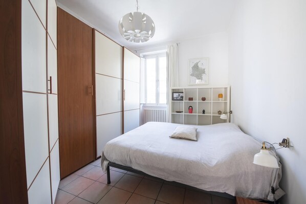 Quarto com cama, guarda-roupa, prateleira com objetos decorativos e luminária suspensa.