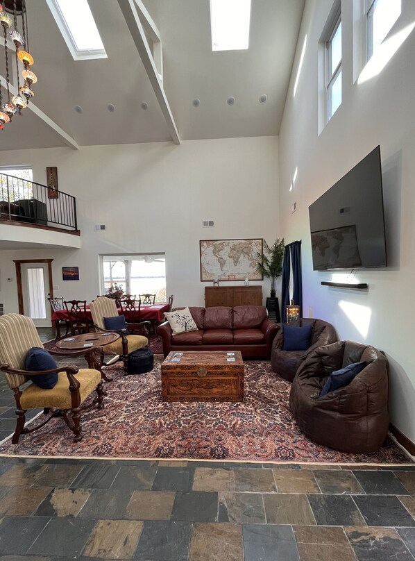 A spacious living room with a high ceiling, a large world map on the wall, a flat-screen TV, and a cozy seating area with a patterned rug and leather sofas. - WHISPERING WINDS! Hot Tub and Pool. (Hernando)