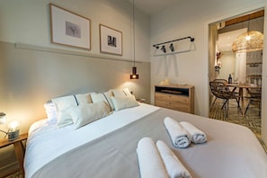 A hotel room with a neatly made bed, a side table with a lamp, a wooden dresser, a dining area with chairs, and two framed pictures on the wall.