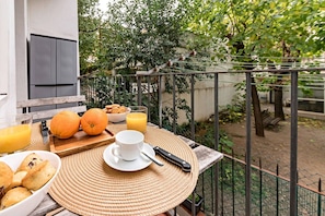 A breakfast setup with orange juice, orange, and pastries on a balcony with a view of trees and a fence.