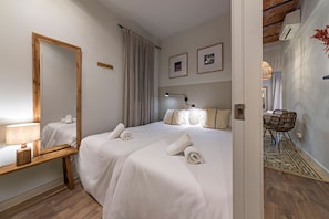 A hotel room with two beds, a wooden nightstand, a mirror, a desk with a chair, and a dining area with a table and chairs.