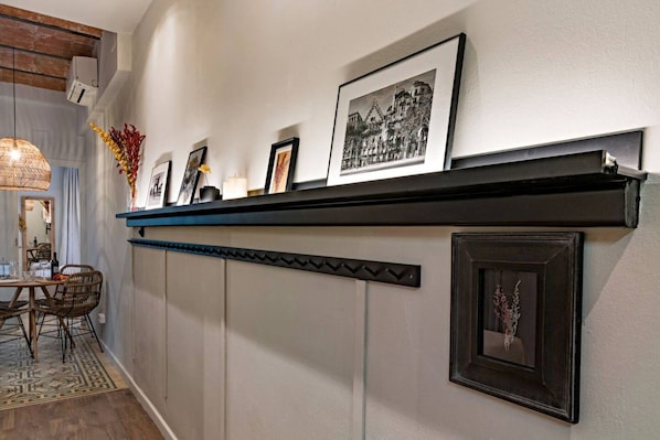 A hallway with a black shelf displaying framed pictures, a dining area with a table and chairs, and a patterned rug on the floor.