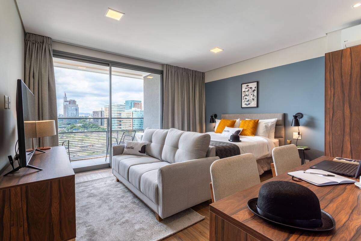 A modern hotel room with a large window, a sofa, a desk with a laptop, and a bed with orange pillows.