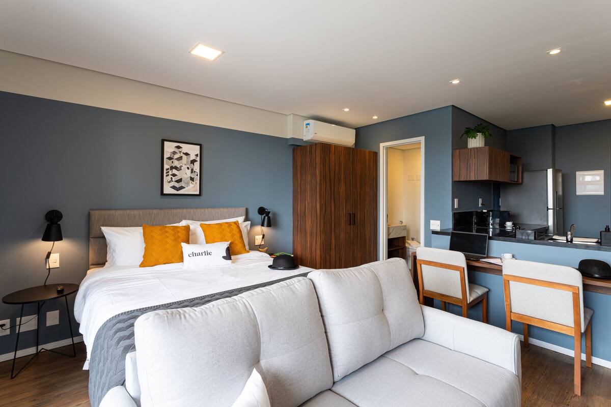 A modern hotel room with a bed, a sofa, a desk, and a kitchenette.