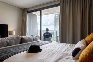 Hotellihuone, jossa on suuri sänky, sohva, taulutelevisio ja parveke, jolta on näkymä kaupunkiin.