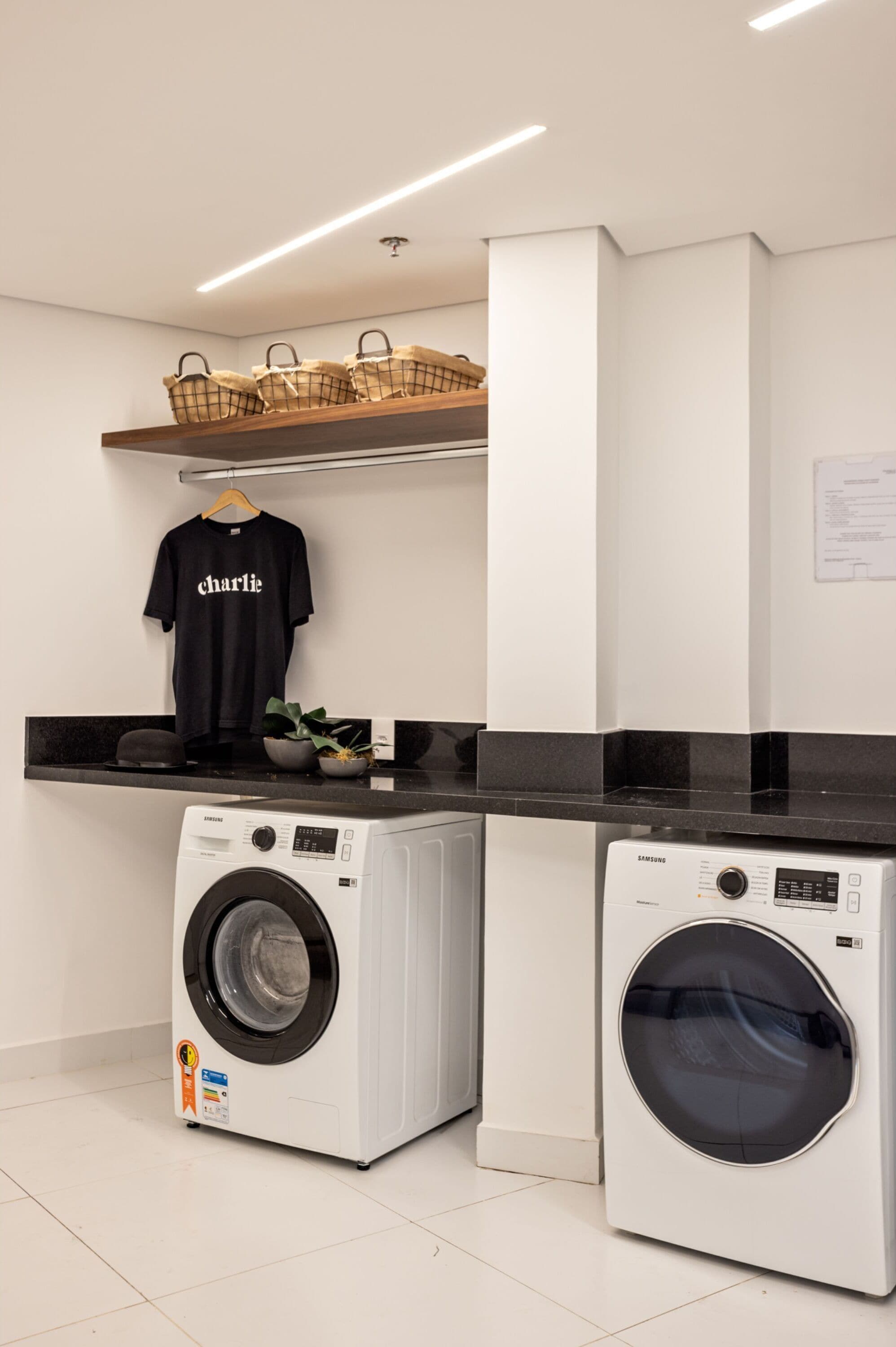 A laundry area with two washing machines, a black t-shirt hanging on a rack, and a shelf with baskets.