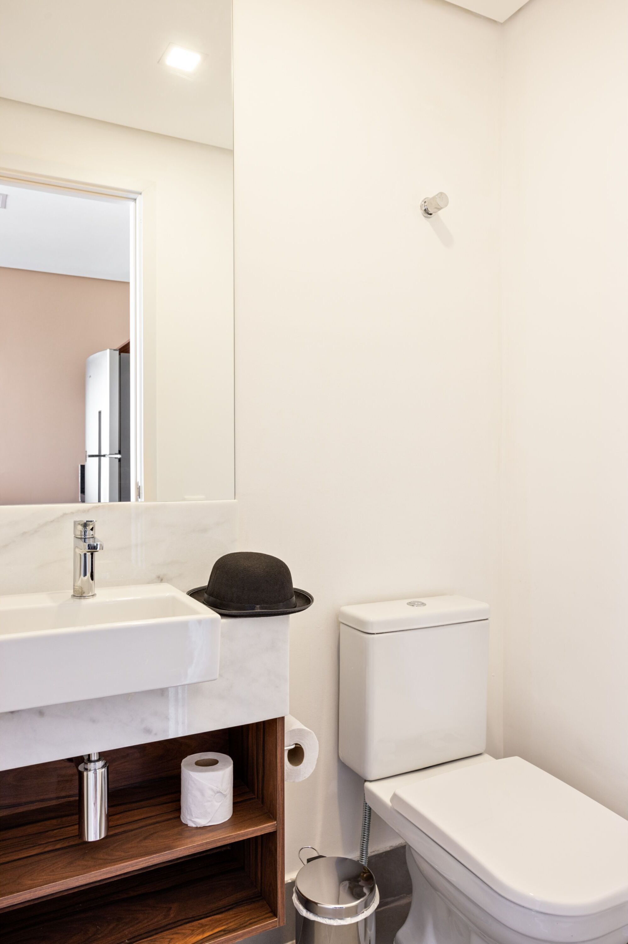 Casa de banho com lavatório branco, espelho, sanita e um chapéu preto no balcão.