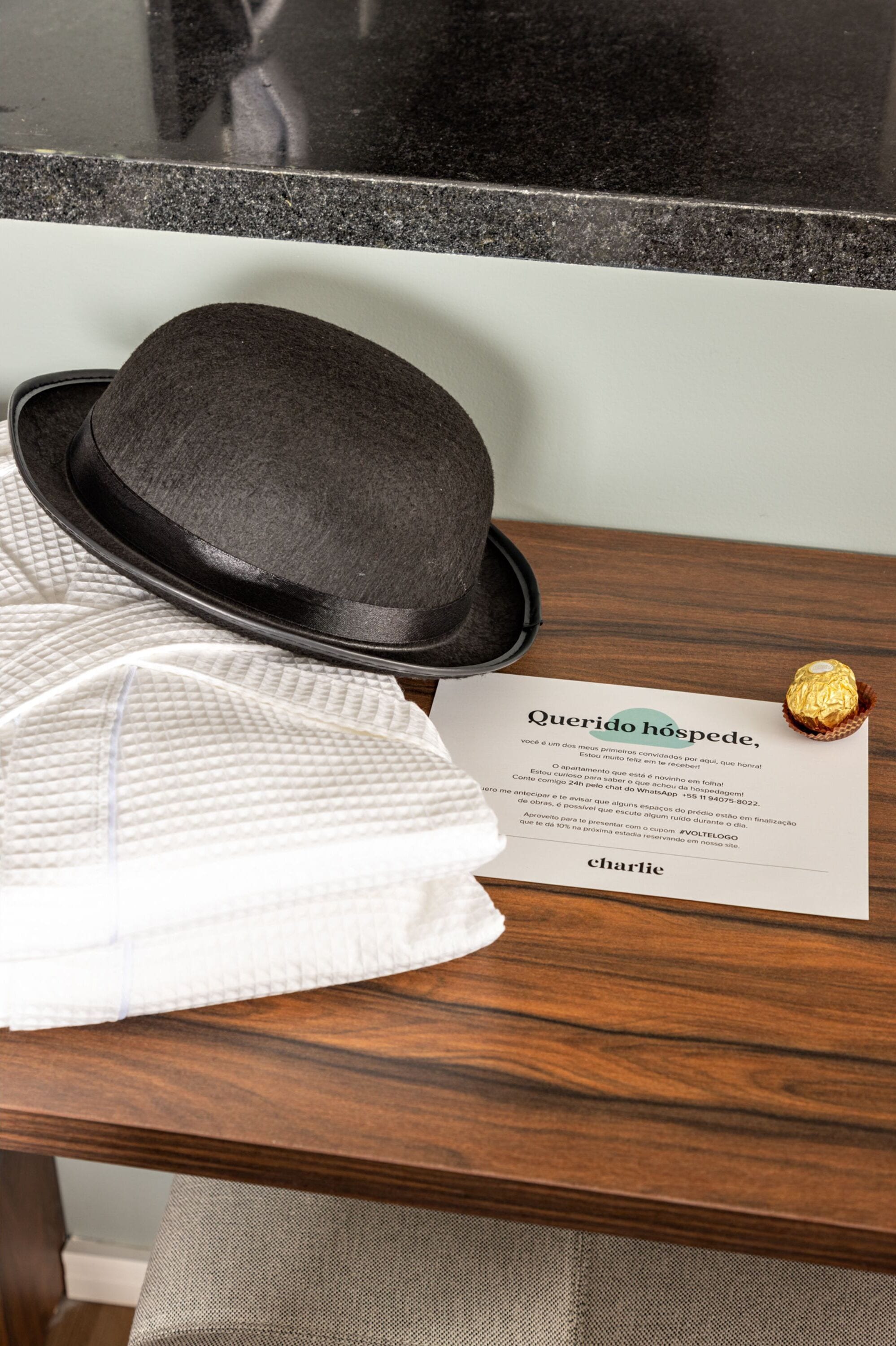 A black hat, a white shirt, a gold-wrapped item, and a "Querido huésped" welcome card on a wooden table.