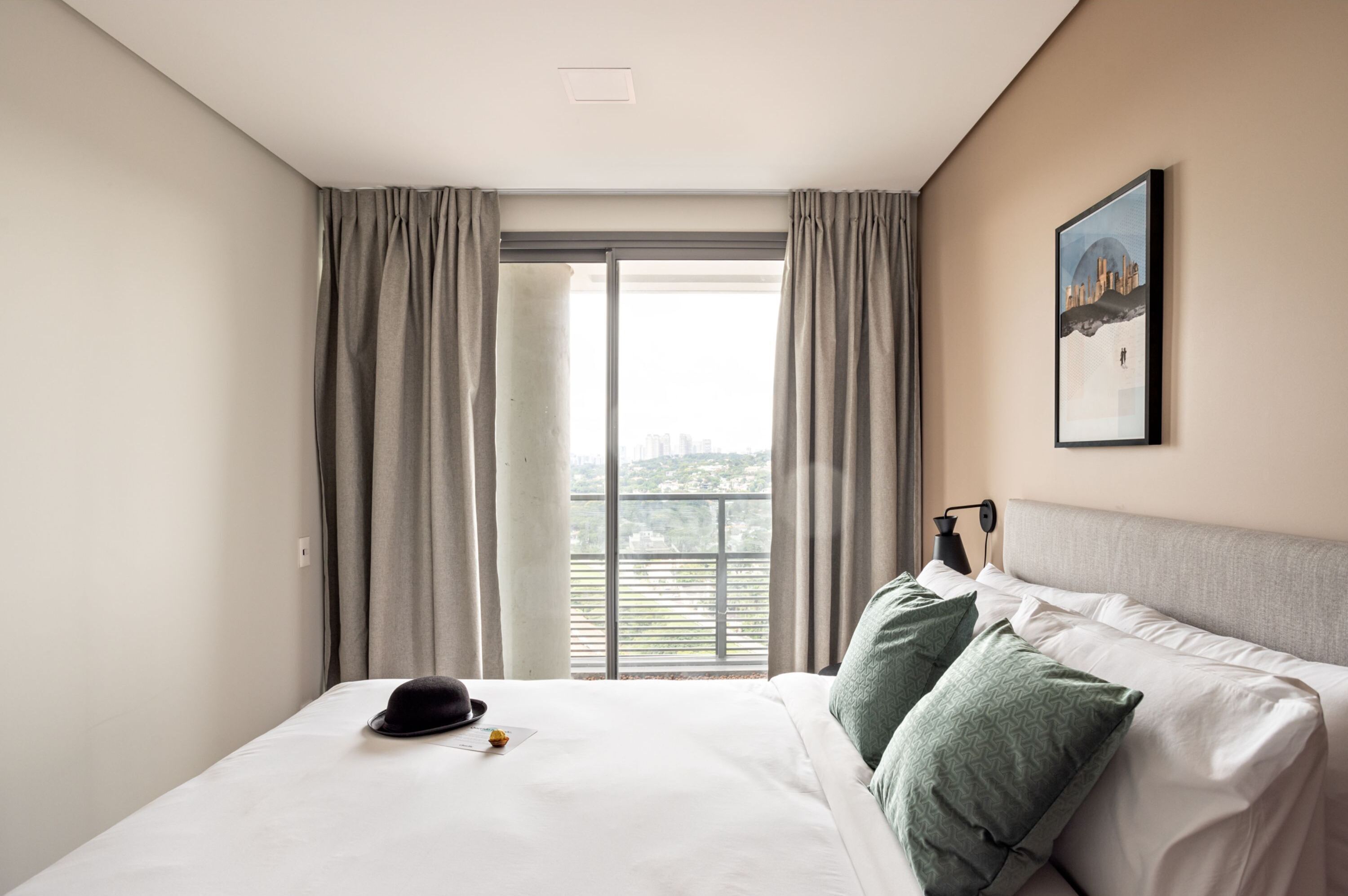 A hotel room with a bed, a window with curtains, a framed picture on the wall, and a hat on the bed.