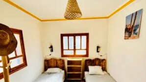Quarto com duas camas de solteiro, piso de madeira, ventilador de teto e janela com moldura de madeira.