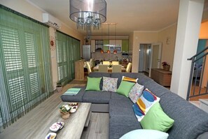 Ein modernes Wohnzimmer mit einem grauen Ecksofa, grünen Kissen und einem Holztisch. Im Hintergrund ist eine Küche mit Edelstahlgeräten zu sehen.