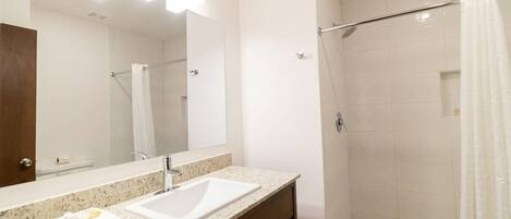 Un baño moderno con encimera de granito, un lavamanos blanco, un espejo grande y una ducha a ras de suelo con puerta de vidrio.