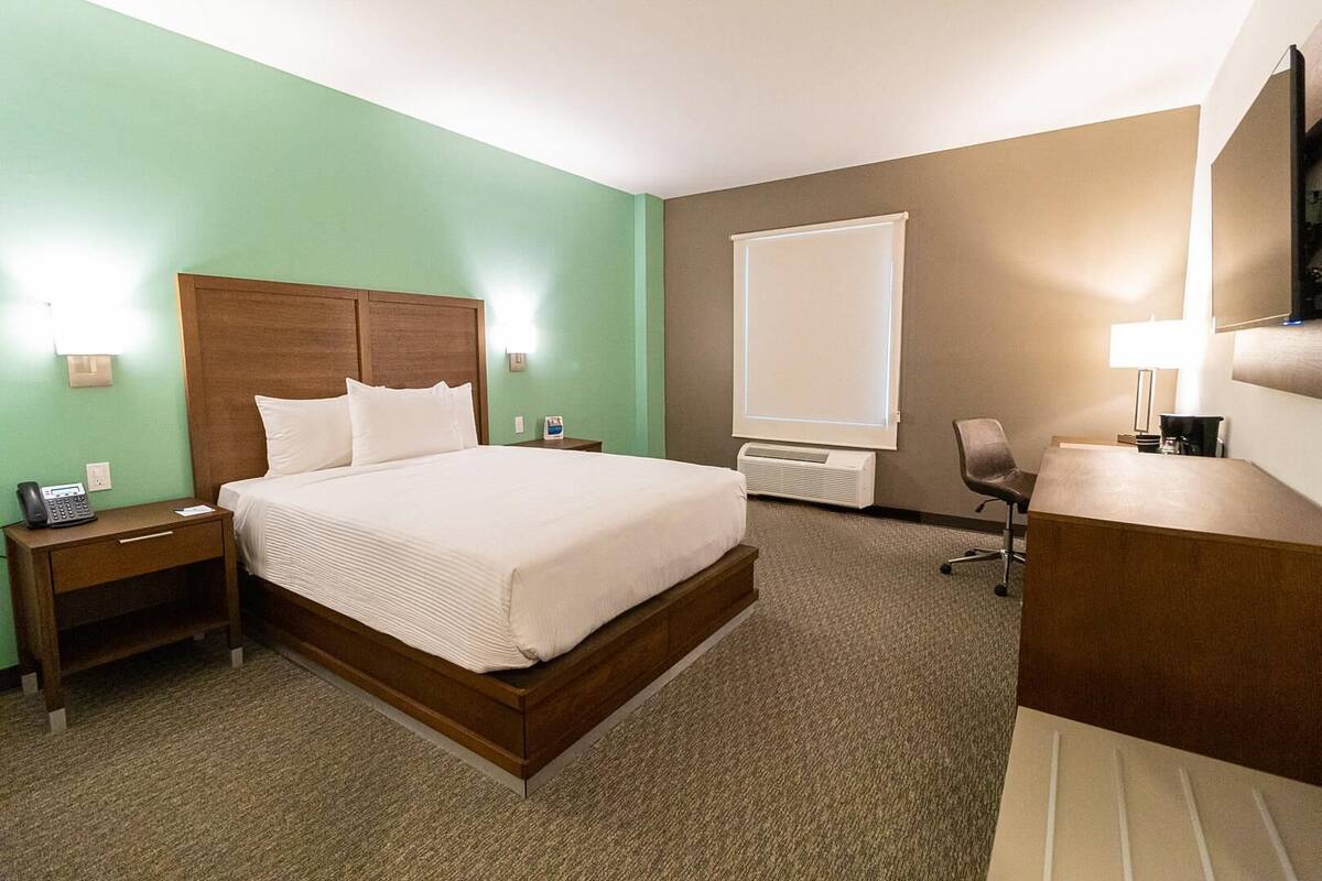 A hotel room with a bed, bedside table, lamp, desk, chair, and a large window.