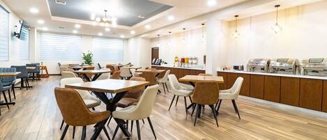 A modern dining area with wooden flooring, multiple tables and chairs, a kitchen counter with food dispensers, and a ceiling with recessed lighting.