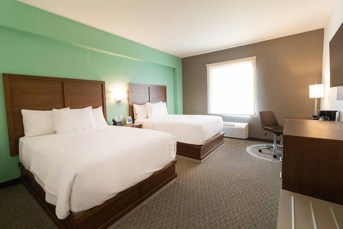 A hotel room with two beds, a desk, a chair, a lamp, and a window with blinds.