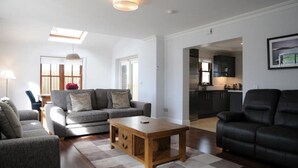 A modern living room with a grey sofa, a wooden coffee table, and a black leather armchair. There is a kitchen visible through an open doorway. - Number Two Dingle: Large Family Holiday Home (Dingle)