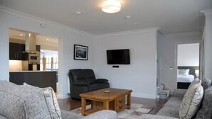 A modern living room with grey sofas, a wooden coffee table, a black leather chair, a flat-screen TV, and a kitchen visible through an open door. - Number Two Dingle: Large Family Holiday Home (Dingle)