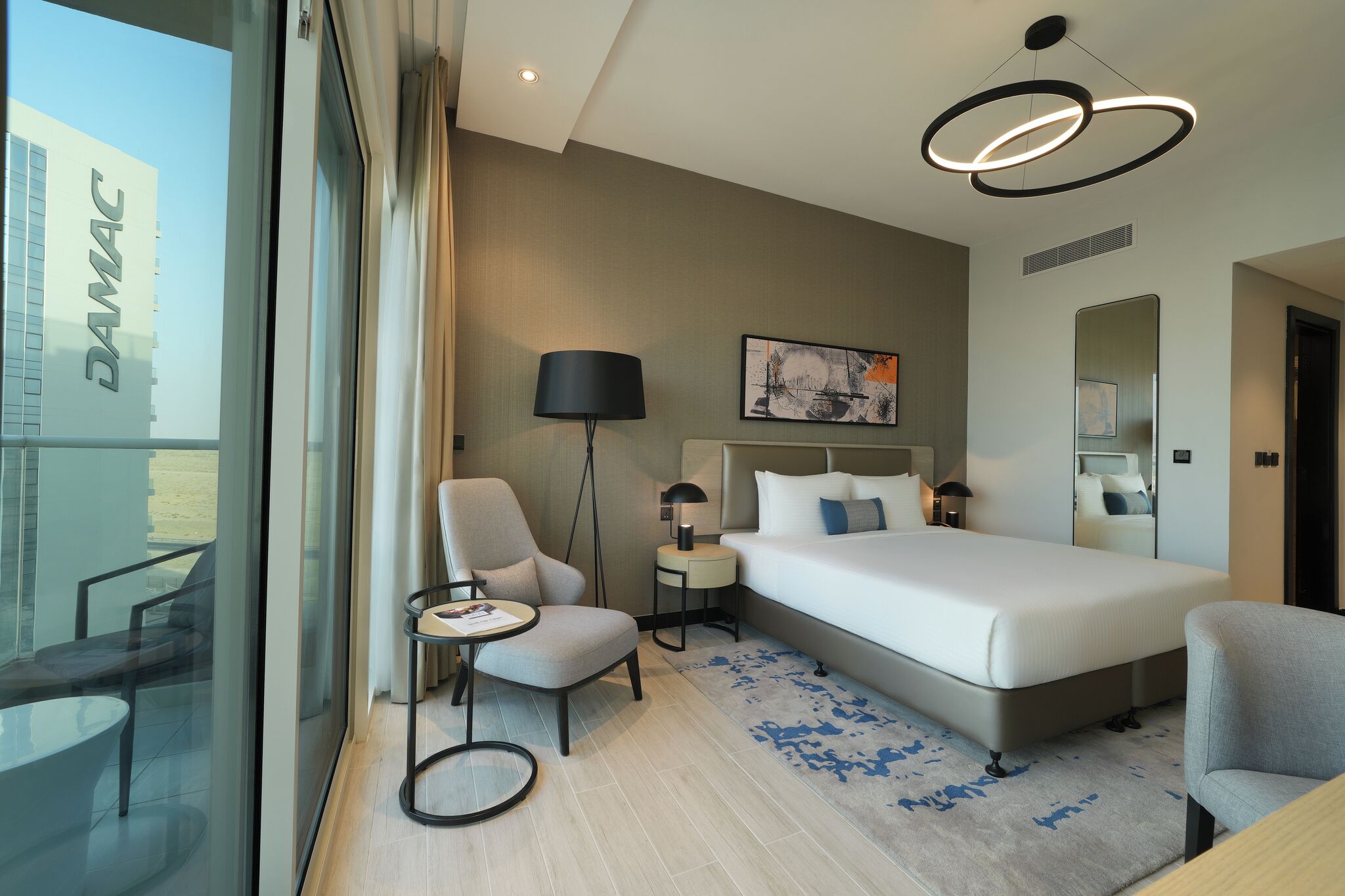 Une chambre d’hôtel moderne avec un grand lit, un bureau, une chaise, une petite table, une lampe et une grande fenêtre.
