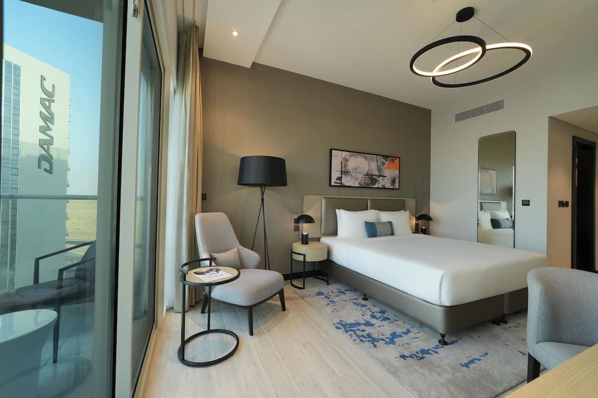Une chambre d’hôtel moderne avec un grand lit, un bureau, une chaise, une petite table, une lampe et une grande fenêtre.