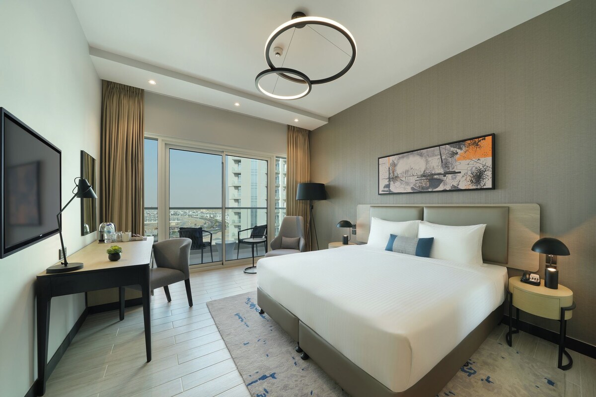Quarto de hotel com cama, escrivaninha, cadeira, mesas de cabeceira e televisão.