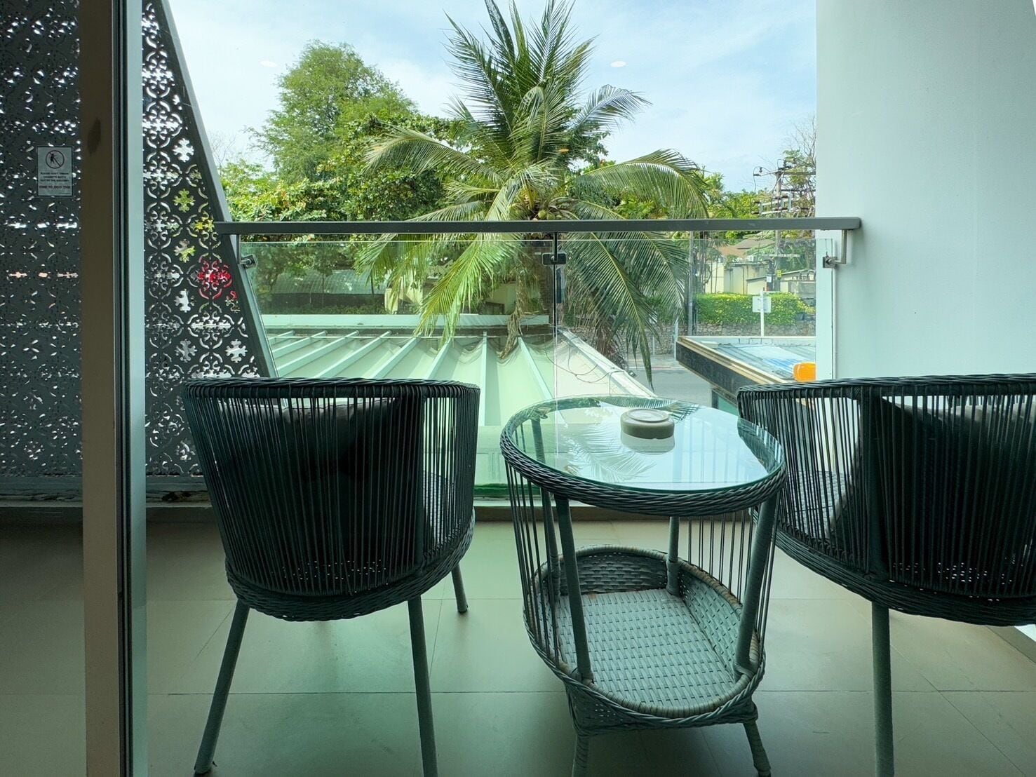 A balcony with two wicker chairs and a small table, overlooking a green area with trees and a building in the background.