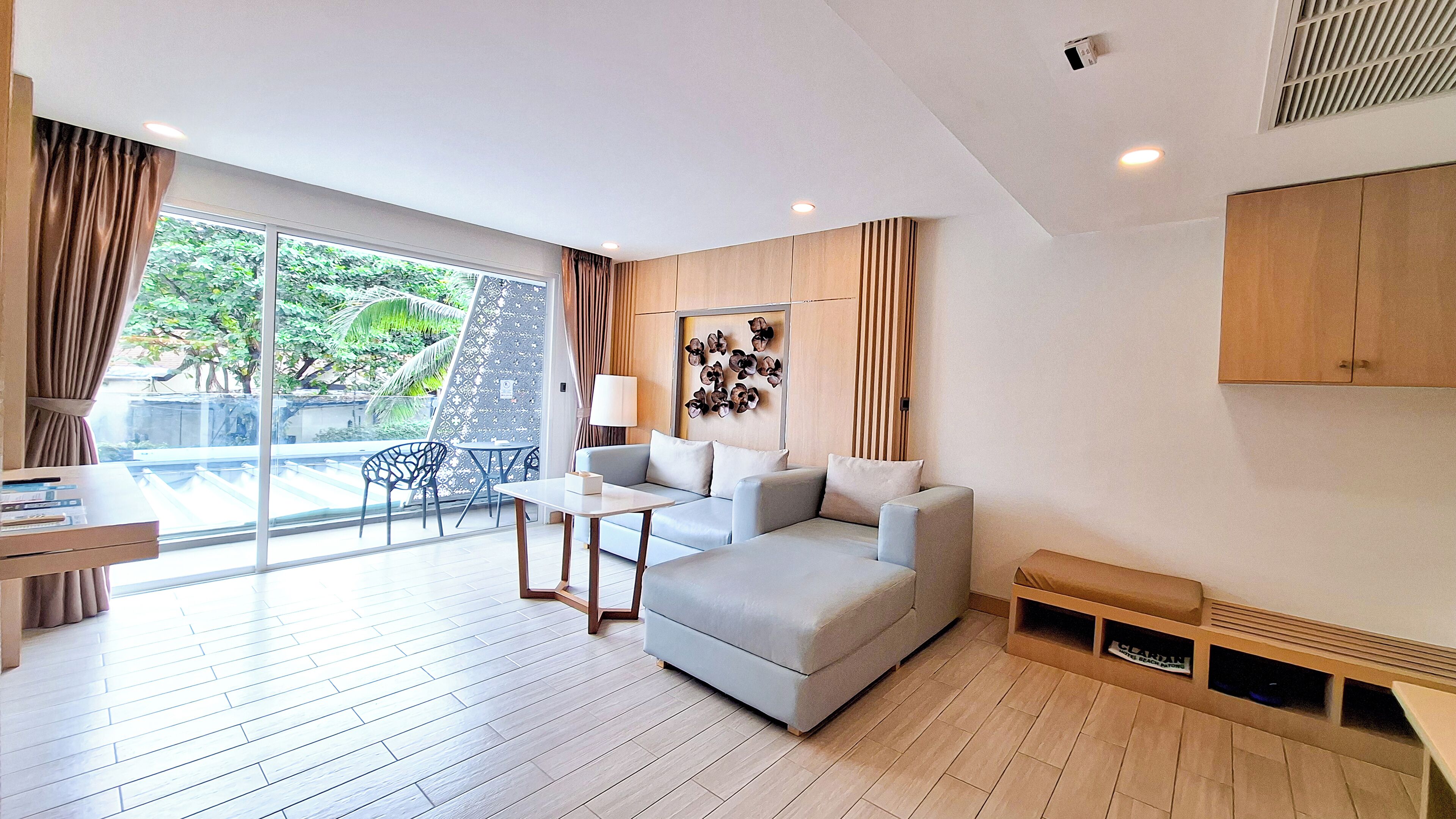 A modern living room with a sofa, a small table, and a lamp. There is a large window with curtains and a view of greenery outside.