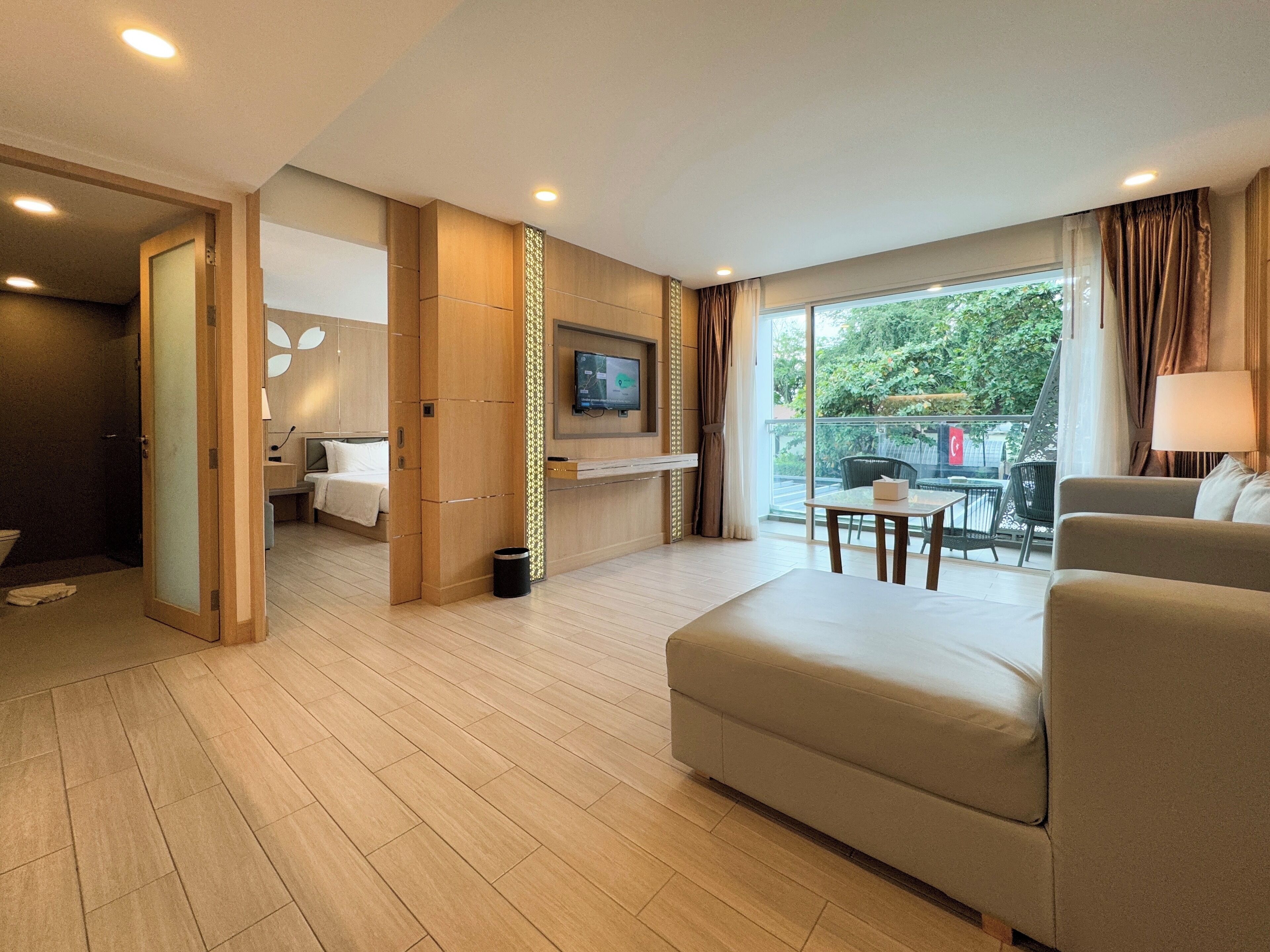 A modern hotel room with a large bed, a TV mounted on the wall, a balcony with a table and chairs, and a wooden floor.