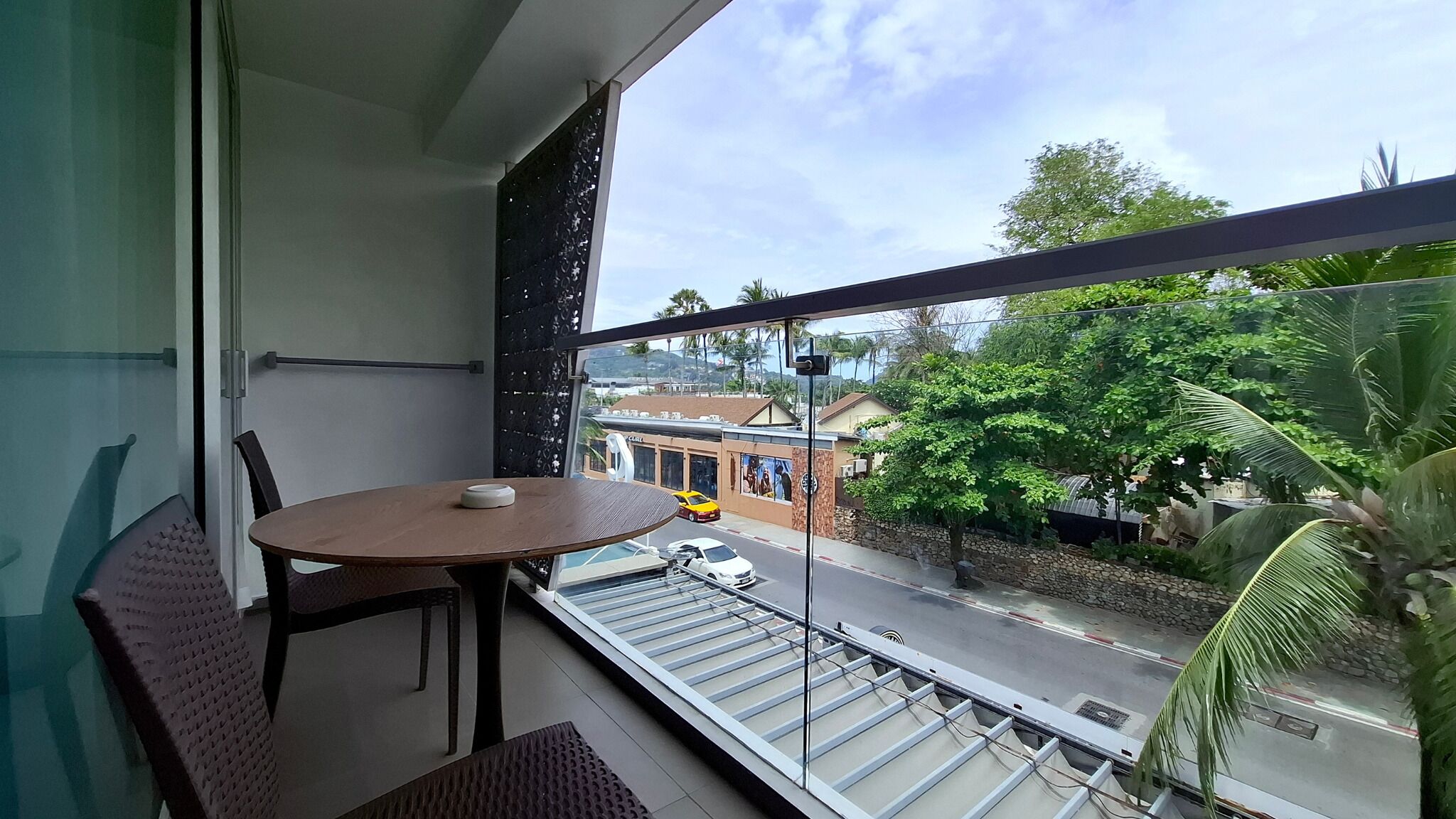 A balcony with a table and chairs, a view of a street with cars and buildings, and a large window with a view of greenery and a clear sky.