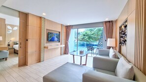 A modern hotel room with a flat-screen TV, a sofa, a dining table, and a balcony with a view of greenery. - Clarian Hotel Beach Patong (Patong)