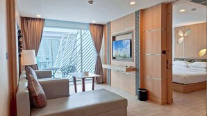 A modern hotel room with a sofa, a small table, a TV mounted on the wall, a bed with a headboard, and a large window with curtains. - Clarian Hotel Beach Patong (Patong)