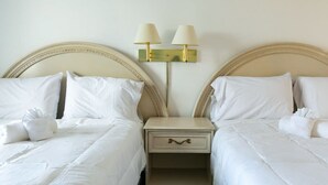 Habitación de hotel con dos camas, cada una con sábanas y almohadas blancas, una mesita de noche con lámpara y una lámpara de pared.