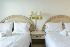 Habitación de hotel con dos camas, cada una con sábanas y almohadas blancas, una mesita de noche con lámpara y una lámpara de pared.