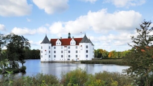 Un edificio grande y blanco con techo rojo, situado junto a un lago tranquilo, rodeado de vegetación.