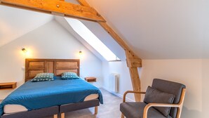 Ein Schlafzimmer mit schrägen Wänden, einem Bett mit blauer Bettdecke, zwei Nachttischen, einem Sessel und einem Holzbalkenbau.