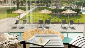 A poolside area with lounge chairs and umbrellas, a magazine on a table, and a view of the pool and garden through a glass window. - Akti Coast Club (Kos)
