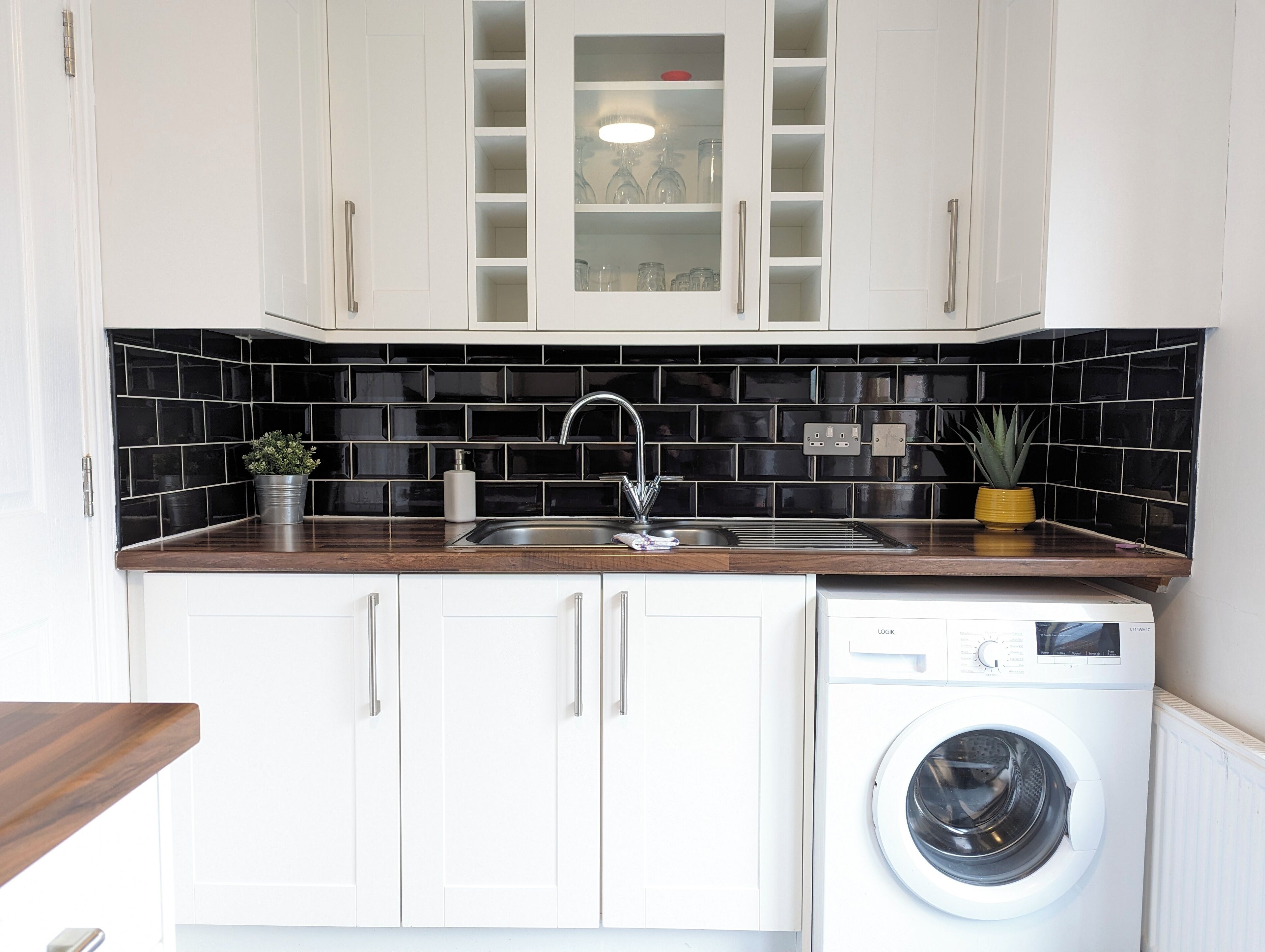 A kitchen with white cabinets, a black tiled backsplash, a stainless steel sink, a white washing machine, and a wooden countertop.