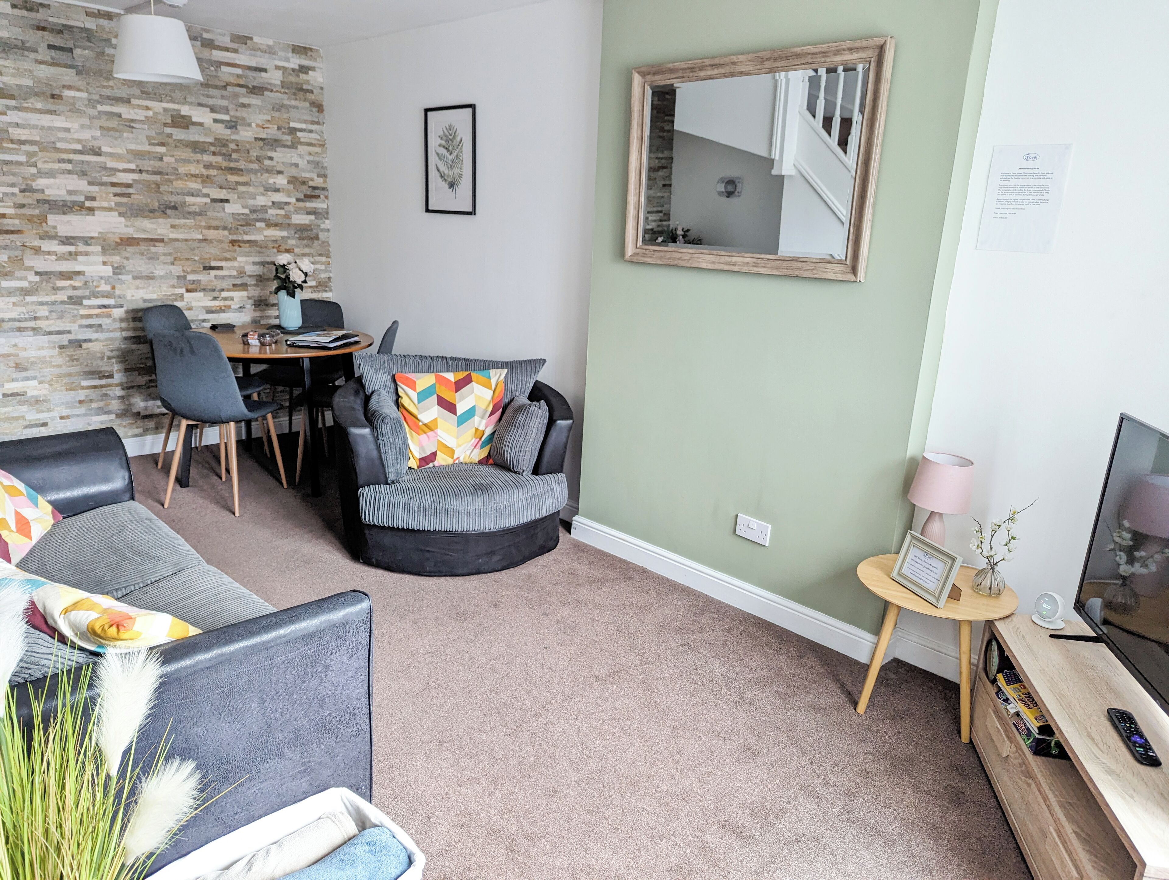 Ein Wohnzimmer mit Steinwand, einem runden schwarzen Sessel mit farbigem Kissen, einem grauen Sofa, einem Holz-TV-Möbel und einem kleinen runden Tisch mit Lampe und gerahmtem Bild.
