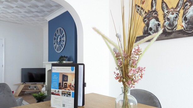 Una stanza con un tavolo in legno, un vaso con piante essiccate, una sveglia appesa al muro e un quadro con asini.