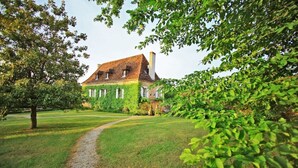 Uma casa grande com telhado inclinado, cercada por um gramado bem cuidado e árvores.