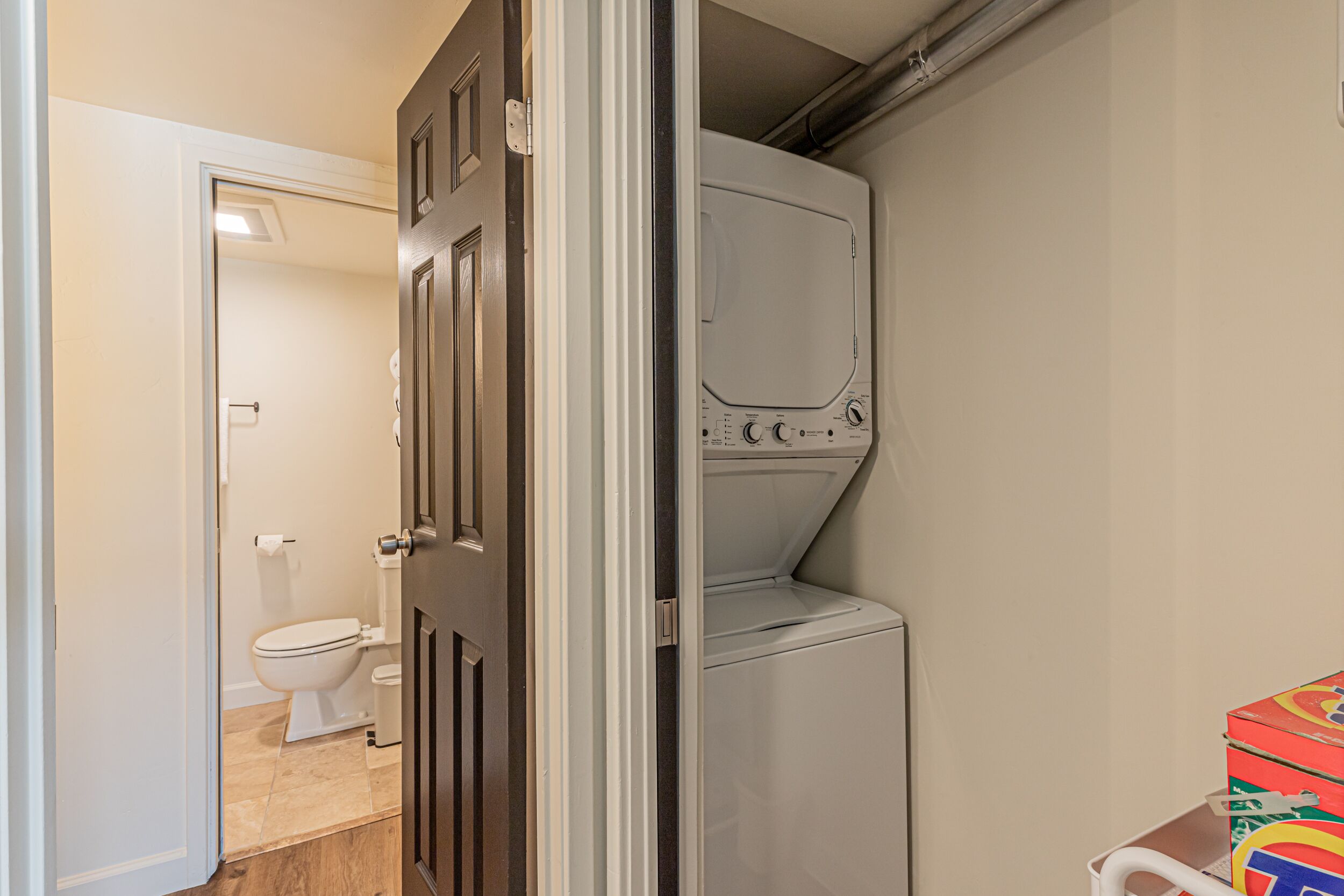 A laundry room with a washing machine and dryer, a toilet, and a door leading to another room.