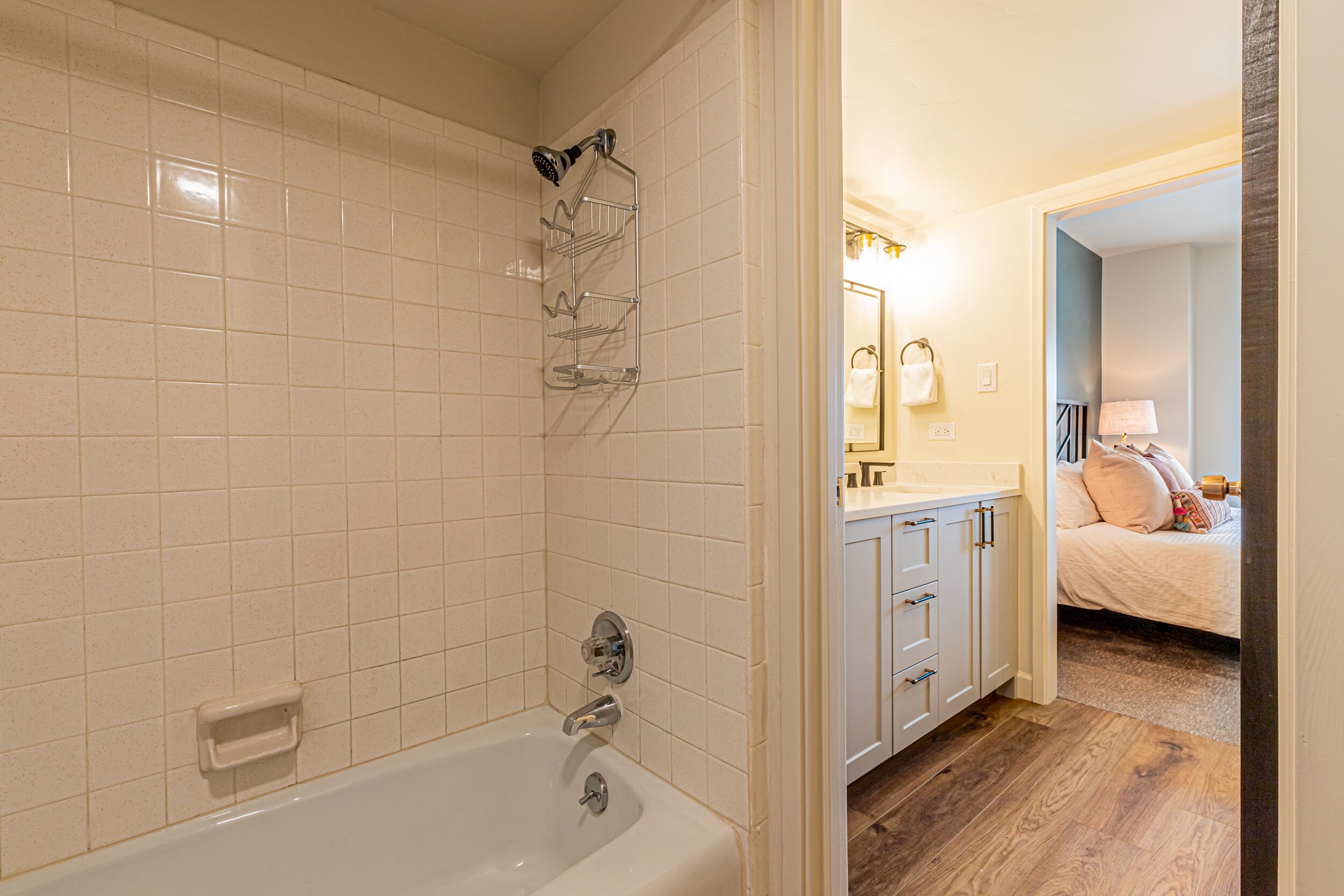 Een badkamer met een bad, douche en toilet. Er is een wastafel met spiegel en een handdoekhouder. De kamer heeft houten vloeren en een deur die naar een slaapkamer leidt.