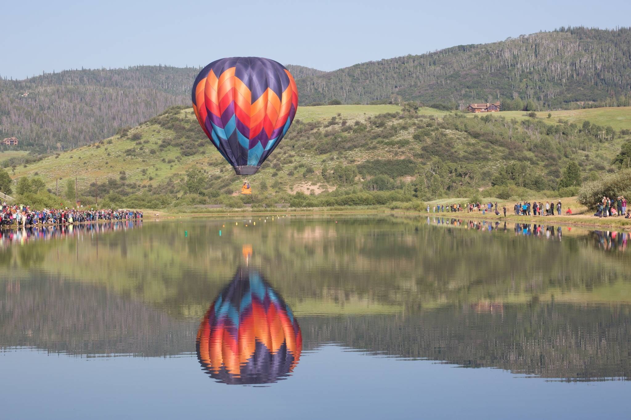 A hot air balloon over a lake with a crowd of people on the shore.