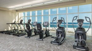 A gym with various exercise machines, large windows, and a view of the beach.