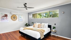 Une chambre moderne avec un grand lit, un ventilateur de plafond, une fenêtre donnant sur des arbres et des œuvres d’art murales.