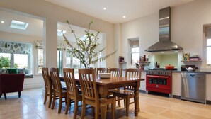 A modern kitchen with a dining area, featuring a central wooden table, chairs, a red oven, and a stainless steel dishwasher.