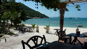 Ein Restaurant direkt am Strand mit Terrassensitzgelegenheiten, mit Blick auf ein ruhiges Meer und eine gebirgige Landschaft.