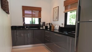 Eine moderne Küche mit dunklen Schränken, einer schwarzen Arbeitsplatte und einem Kühlschrank. Zwei Fenster mit Jalousien, ein Spülbecken und eine Kaffeemaschine befinden sich auf der Arbeitsfläche.